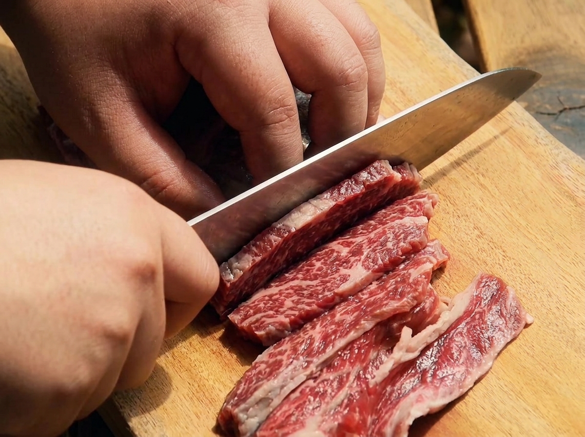Un cuchillo afilado cortando costillas de ternera crudas en tiras finas sobre una tabla de madera.