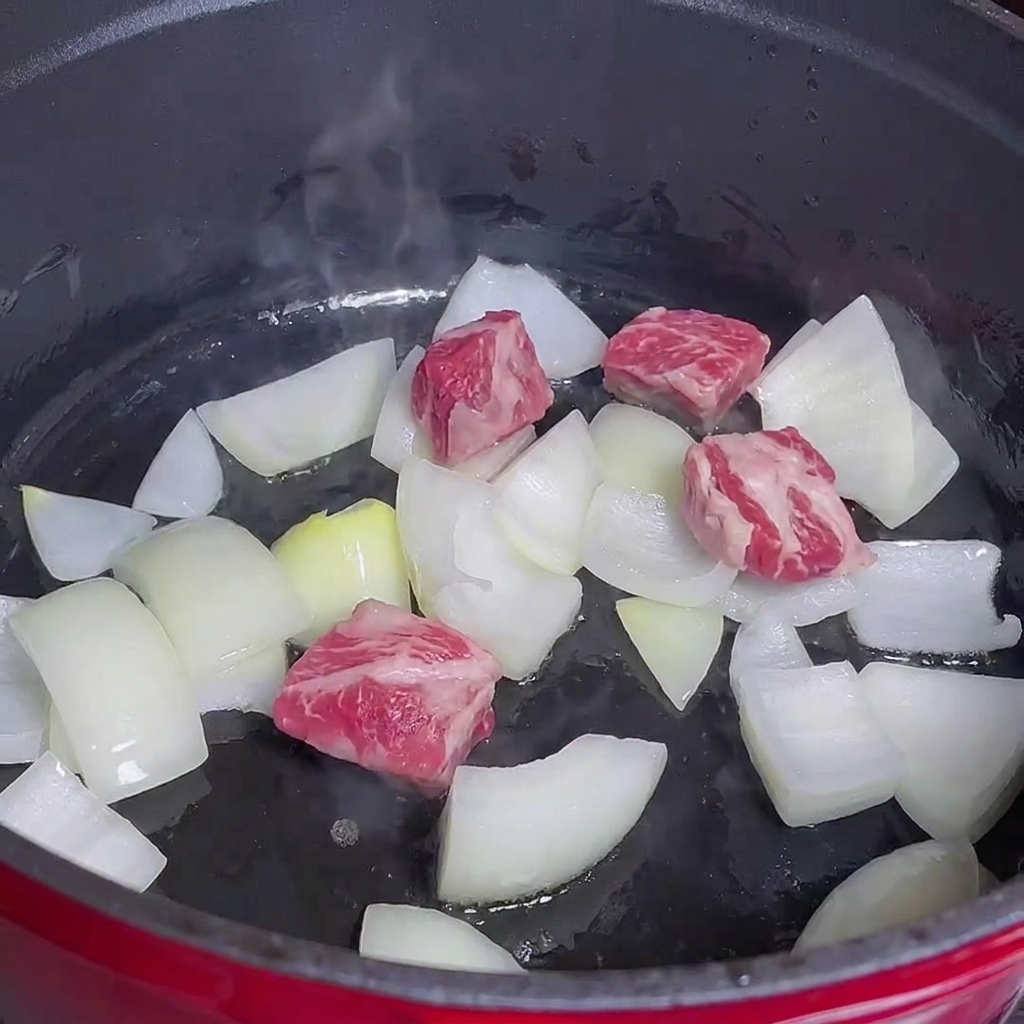 Dados de ternera cocinándose junto a cebollas en una olla roja.