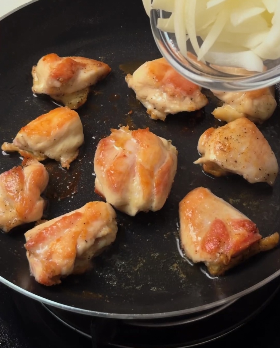 Tiras de cebolla siendo añadidas a la sartén con los trozos de pollo dorados.