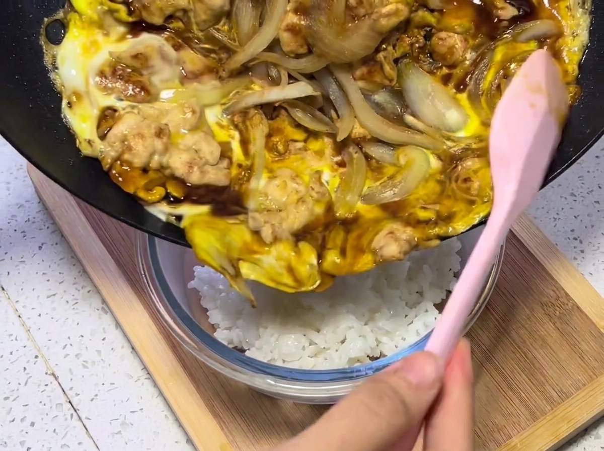 Primer plano de la mezcla de pollo, huevo y cebolla siendo transferida desde la sartén a un bol de arroz blanco humeante.