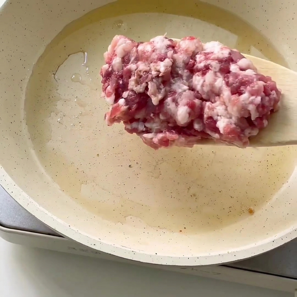 Primer plano de carne picada fresca siendo añadida a una sartén con aceite caliente.