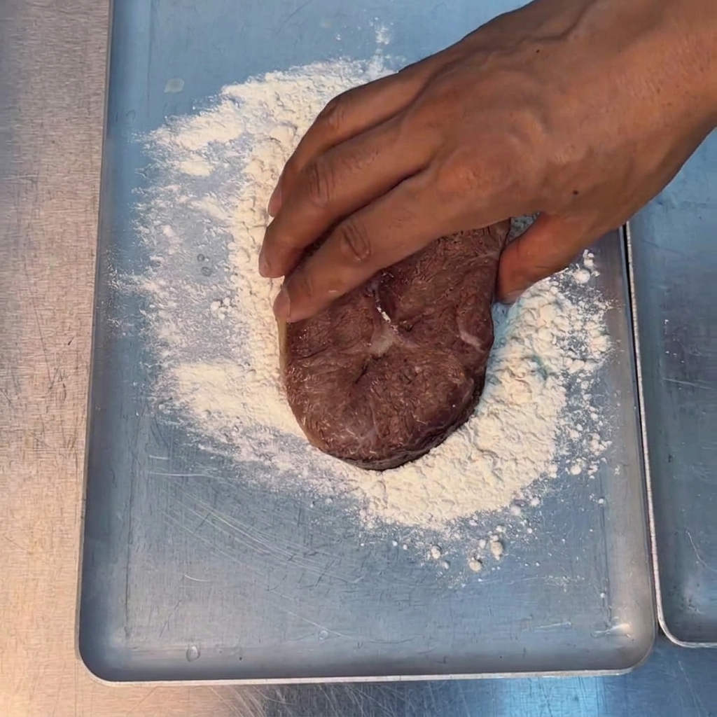 Una mano presionando un trozo grueso de carne sobre harina blanca en una bandeja de metal.
