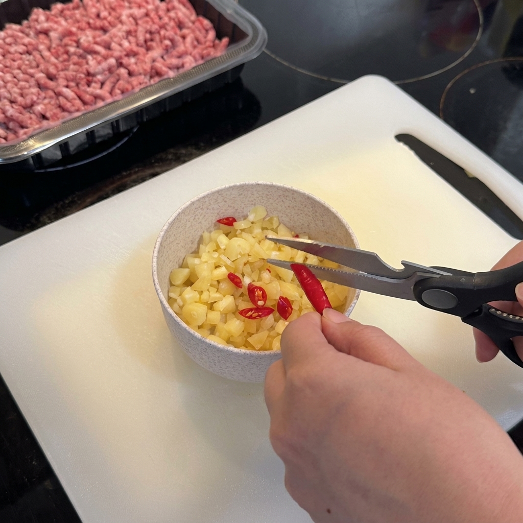 Tijeras de cocina cortando chiles ojo de pájaro rojos vibrantes sobre un tazón que contiene el ajo picado.