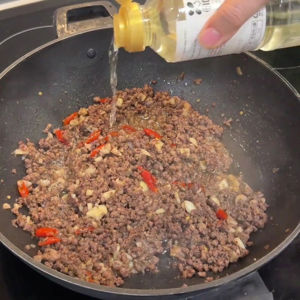 Un chorrito de vino de arroz siendo vertido en un wok que contiene carne picada dorada.