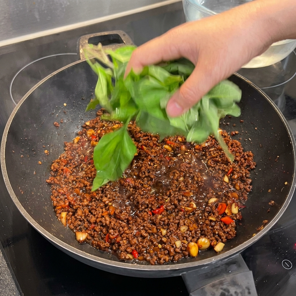 Un gran puñado de hojas de albahaca sagrada fresca siendo añadido a un wok con carne sazonada, listas para marchitarse.