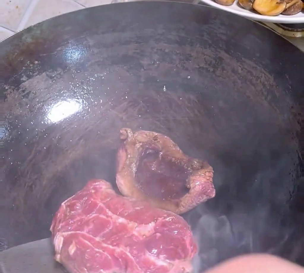 Una espátula de metal dando la vuelta a un trozo de carne a medio dorar dentro de un wok oscuro y caliente.