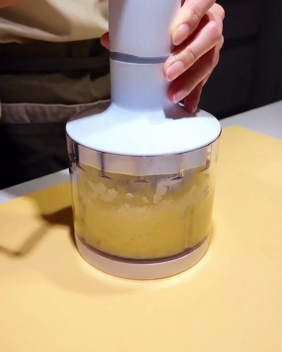 Un pequeño vaso de licuadora lleno con un puré amarillo claro y una base de marinado oscura, listo para mezclarse.