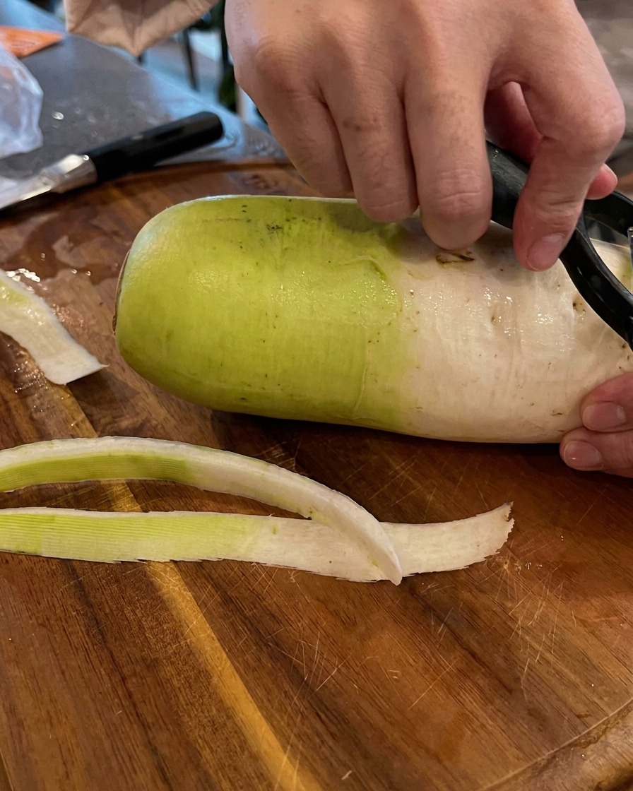 Manos usando un pelador de verduras negro para quitar la piel verde y blanca de un rábano grande sobre una tabla de cortar de madera.