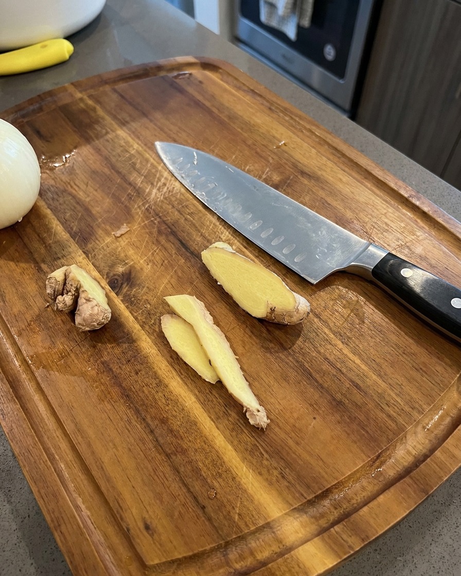 Finas rodajas de jengibre fresco descansando sobre una tabla de cortar de madera junto a un cuchillo de chef y media cebolla.