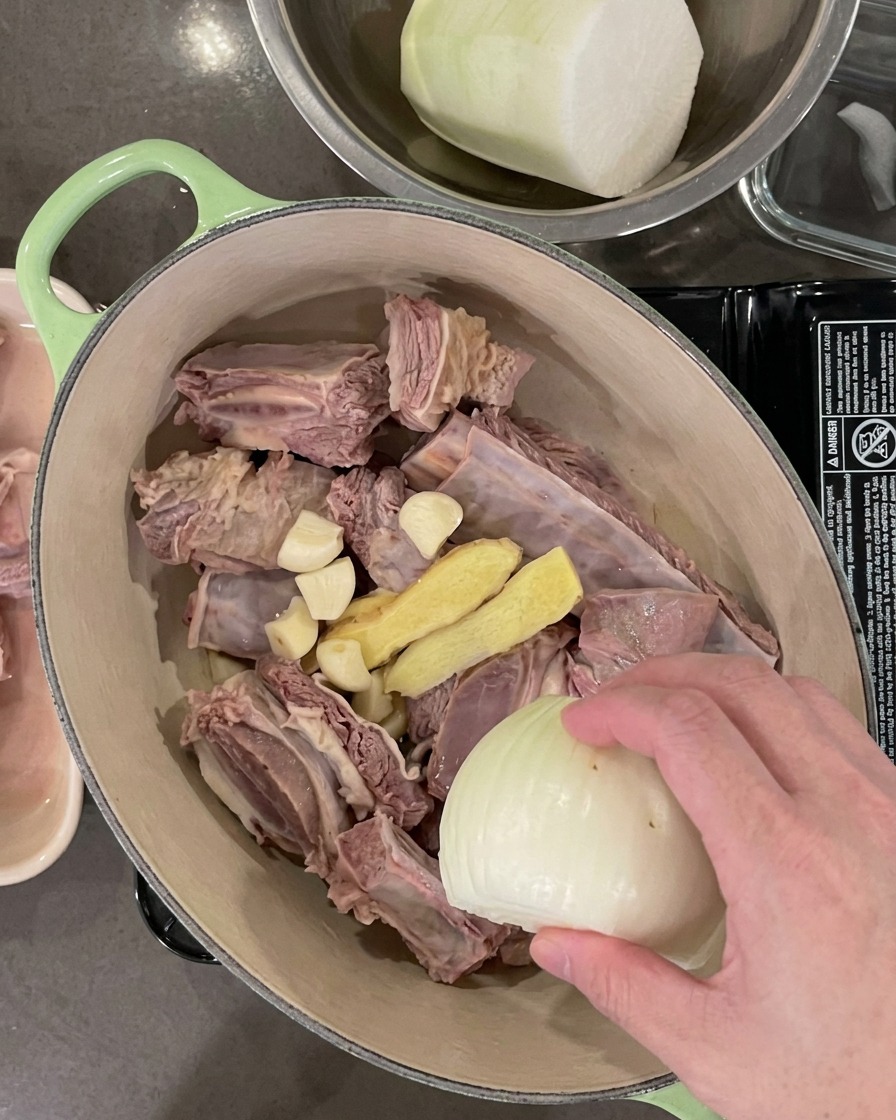 La olla conteniendo las costillas ahora incluye dientes de ajo, rodajas de jengibre, media cebolla y cebollinos.