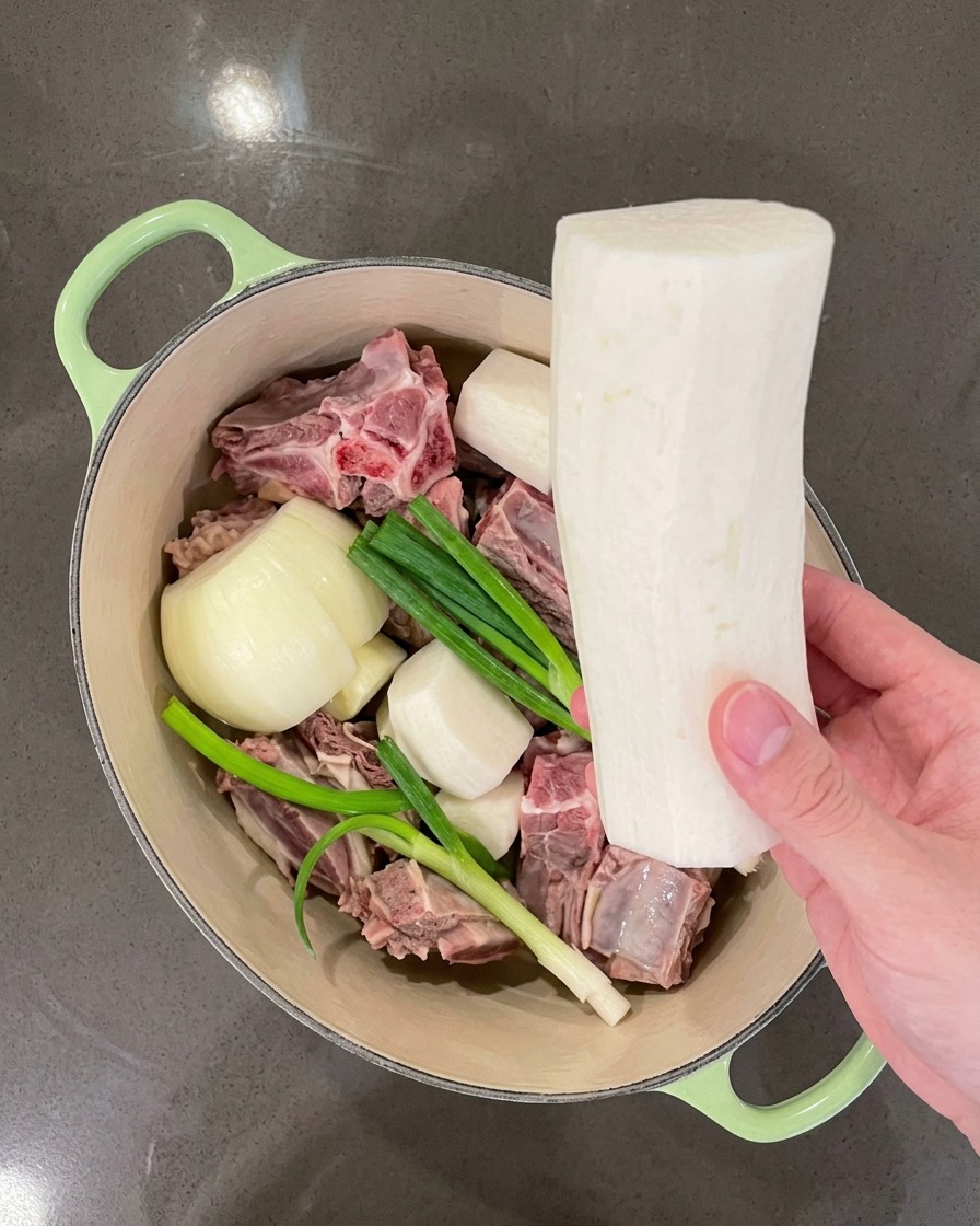 Mano colocando trozos grandes de rábano blanco en la olla junto a la carne y los aromáticos.