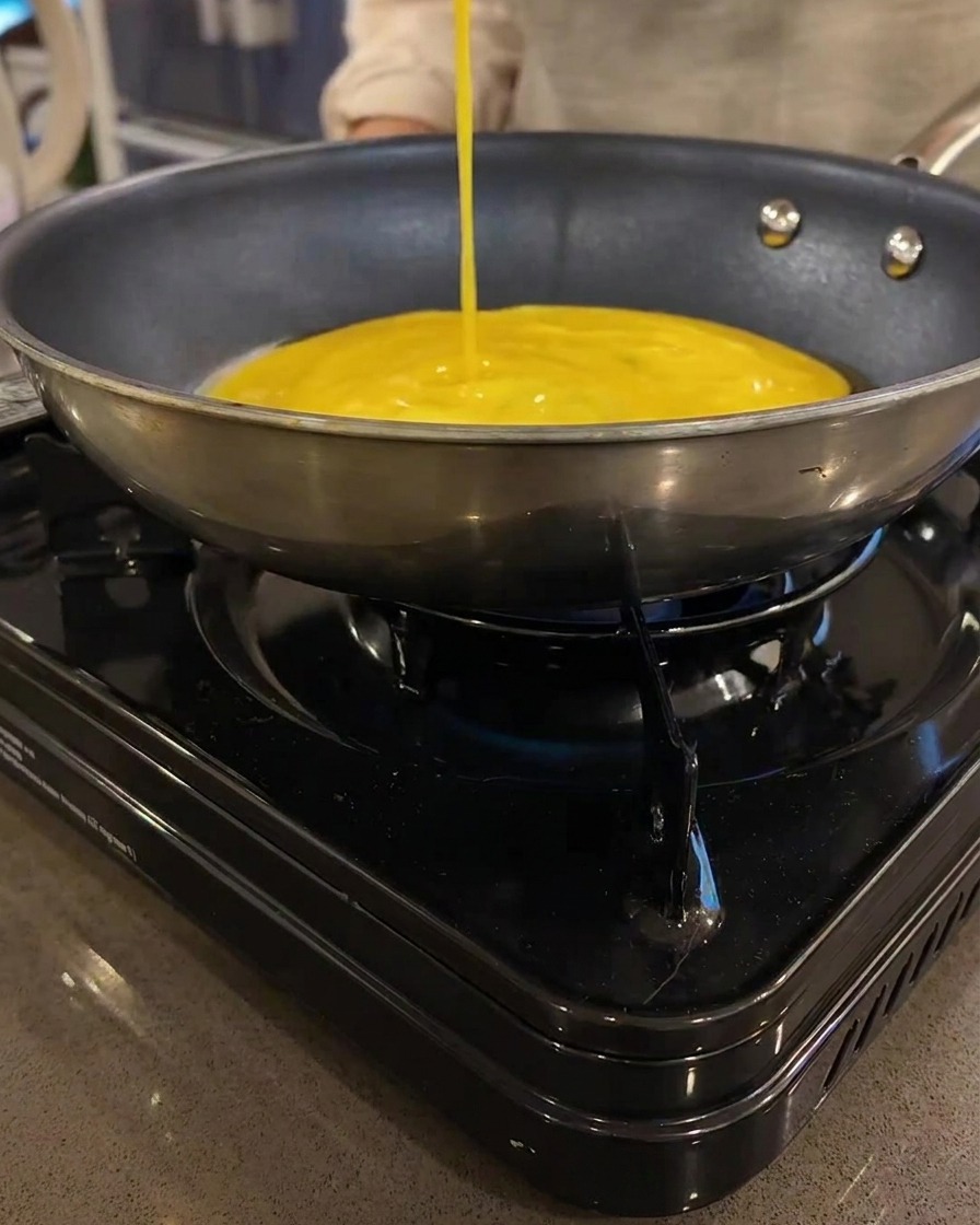 Una fina crepe de huevo amarilla cocinándose en una sartén plana sobre una estufa portátil.