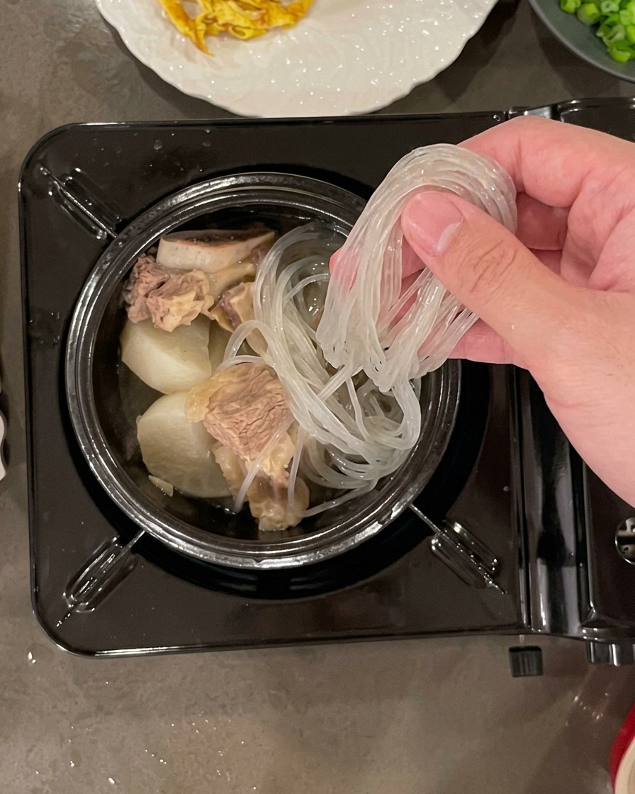 Una mano colocando un manojo de fideos de cristal remojados y translúcidos en una olla de piedra llena de trozos de ternera y rábano.