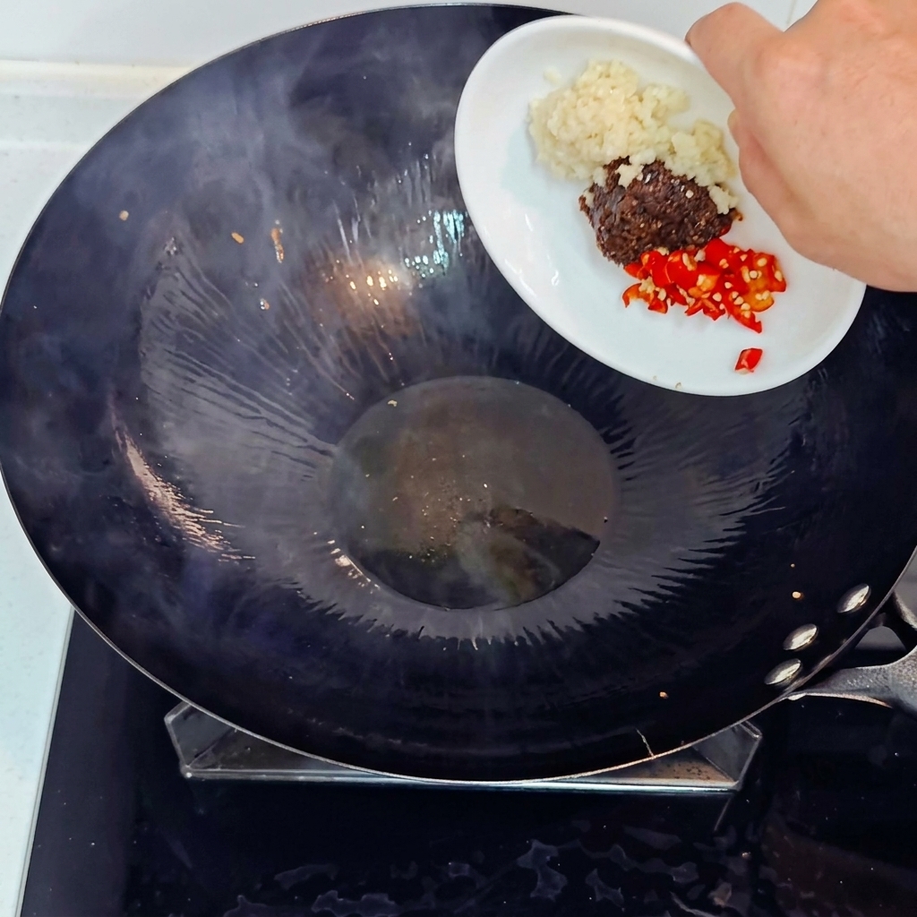 Añadiendo ajo picado, chile rojo troceado y pasta de soja a un wok caliente desde un plato blanco pequeño.