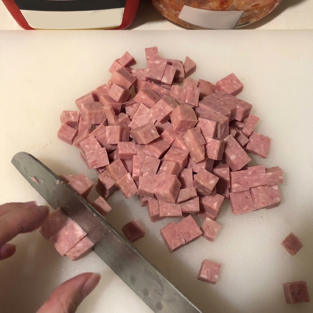 Primer plano del spam siendo cortado en cubos uniformes sobre una superficie blanca, con la hoja de un cuchillo visible.