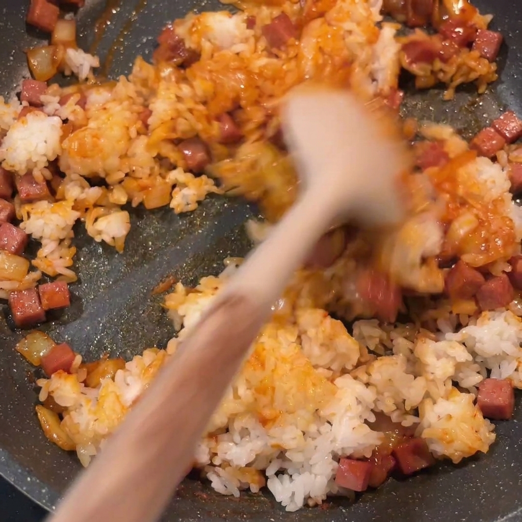 El arroz frito con kimchi terminado siendo removido en una sartén, mostrando un color rojo uniforme y trozos de spam y cebolla bien distribuidos.