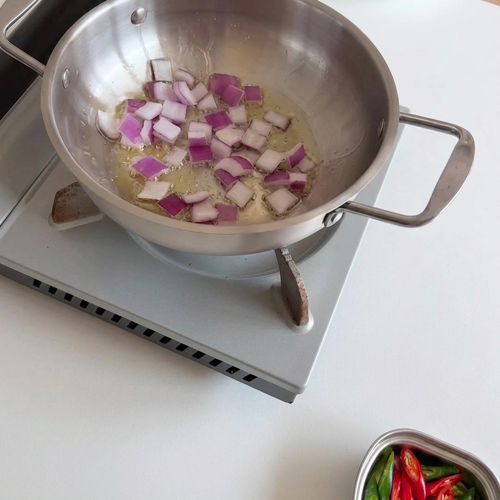 Cebollas moradas picadas chisporroteando y ablandándose en aceite caliente dentro de una sartén de acero inoxidable, siendo revueltas con una cuchara de madera.