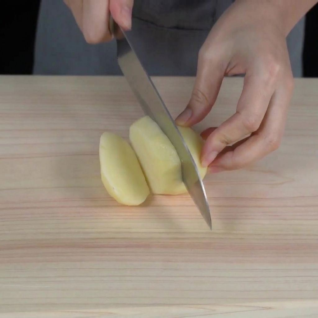 Manos usando un cuchillo de chef para cortar una patata cruda pelada en cubos pequeños sobre una tabla de madera.