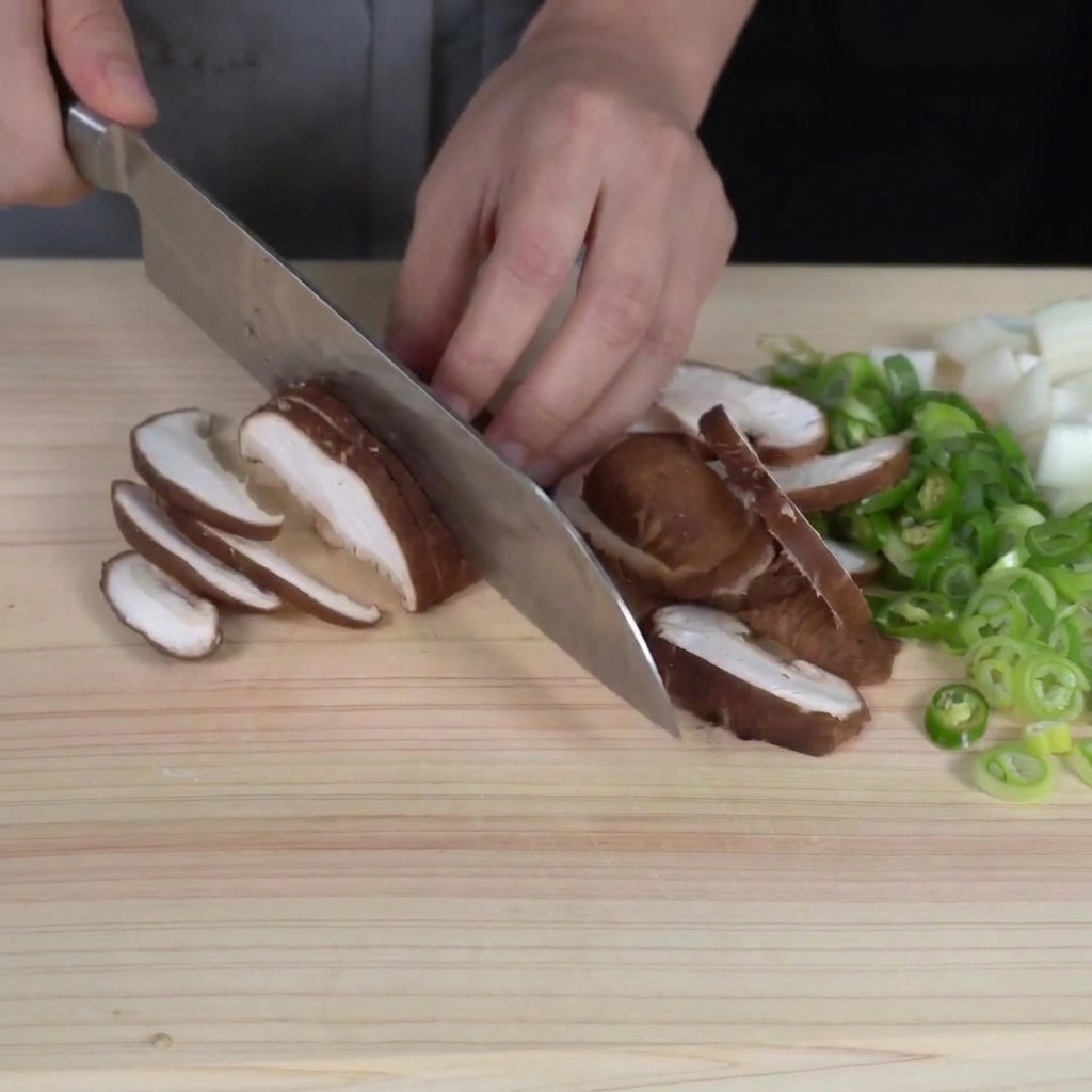 Rebanando finamente setas shiitake frescas sobre una tabla de cortar, con verduras picadas visibles cerca.