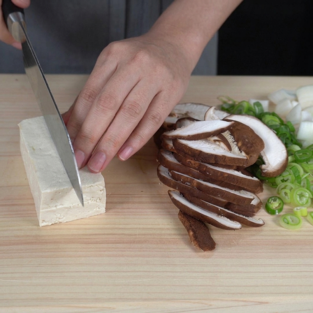 Manos cortando un bloque de tofu firme blanco en cubos uniformes del tamaño de un bocado sobre una tabla de cortar.