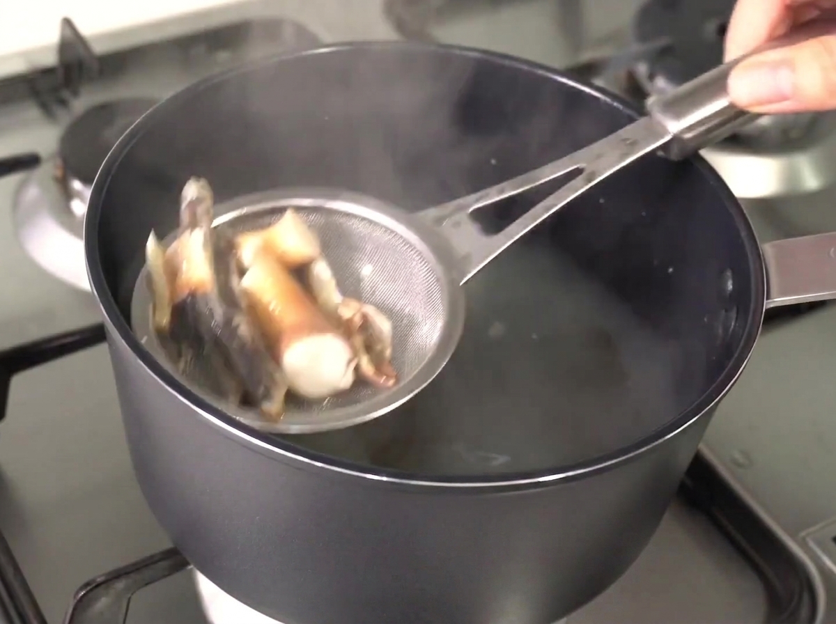 Un colador de metal levantando anchoas y algas ablandadas fuera del caldo caliente y claro.