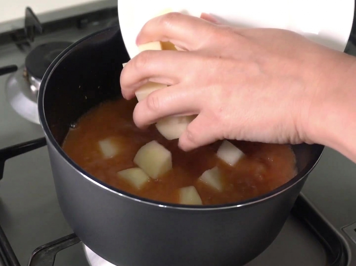 Patatas y cebollas picadas siendo deslizadas desde un plato blanco hacia una olla de caldo rojo hirviendo.
