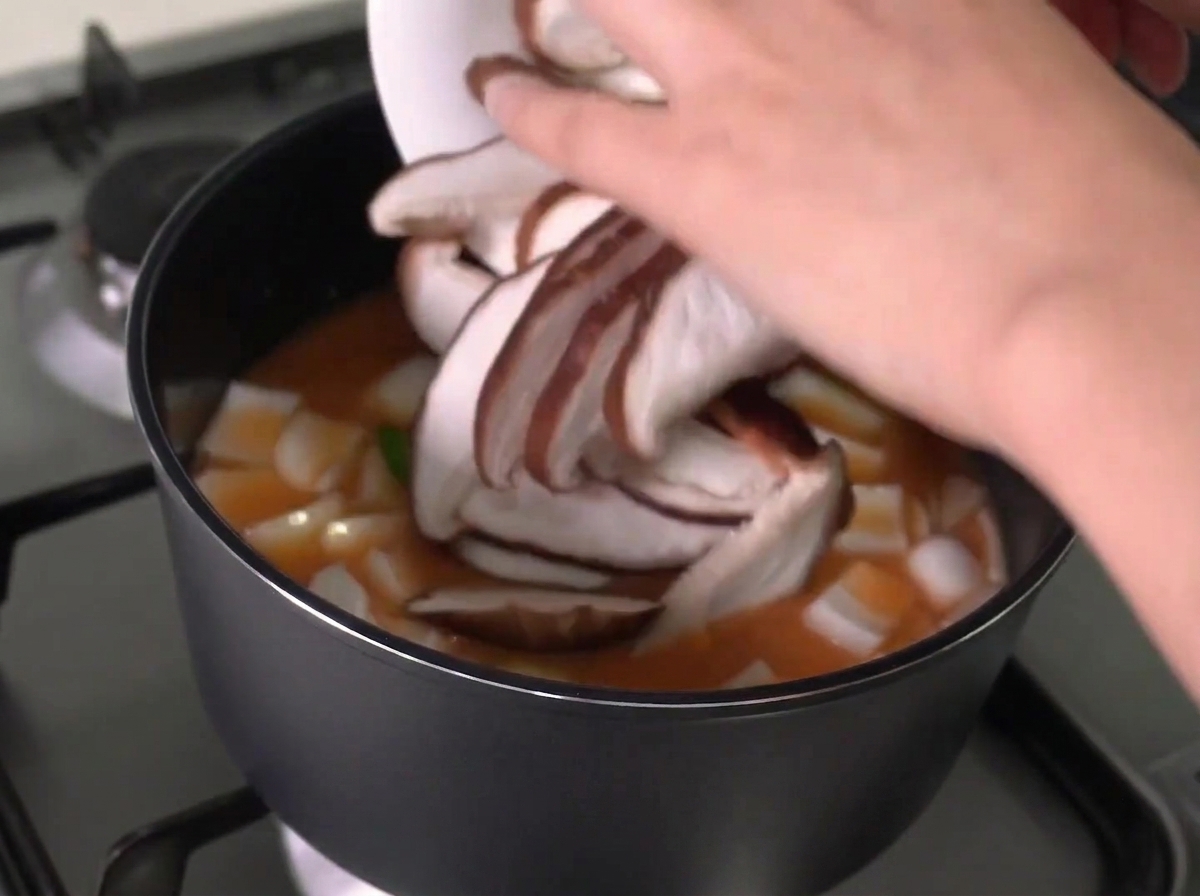 Setas shiitake rebanadas siendo añadidas desde un plato blanco a una olla de guiso hirviendo.