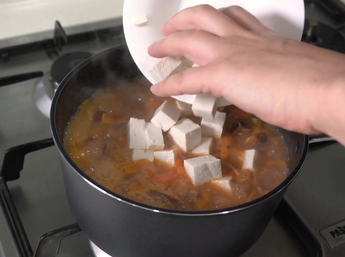 Cubos de tofu fresco siendo añadidos desde un plato blanco a un guiso hirviendo activamente.