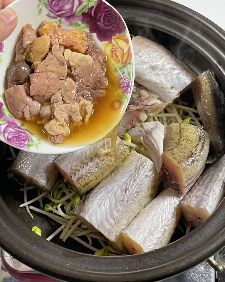 Un pequeño bol floral vertiendo delicadas huevas de abadejo frescas en una olla de barro que ya contiene pescado y brotes de soja.