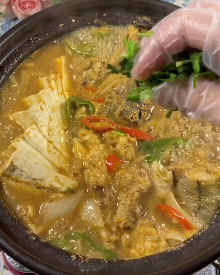 Primer plano de una mano con un guante de plástico transparente colocando cilantro verde fresco en una olla de barro burbujeante con sopa de huevas de abadejo y tofu.