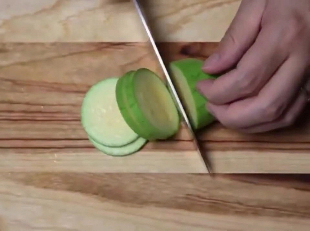 Un calabacín verde siendo cortado en semicírculos uniformes sobre una tabla de madera.
