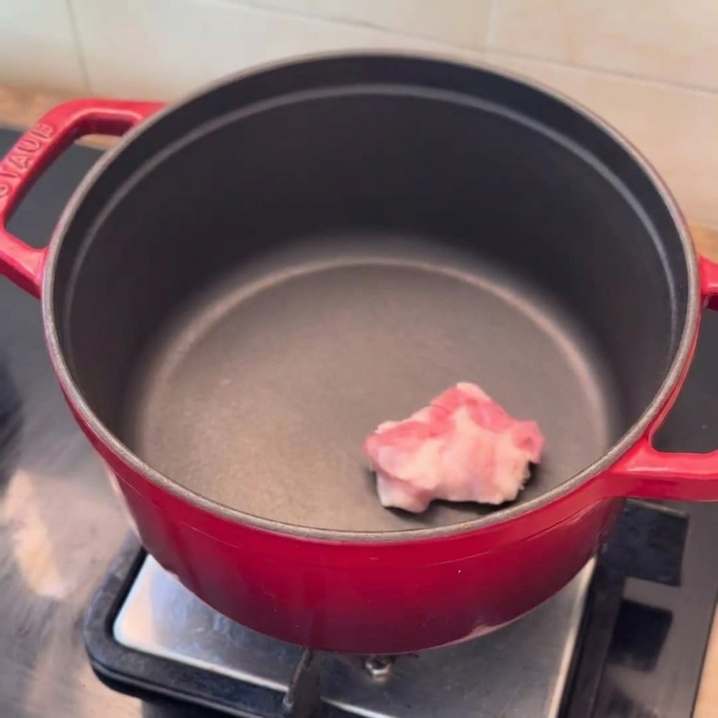 Rodajas de panceta, cuñas de cebolla y ajo picado listos en una olla roja precalentada.