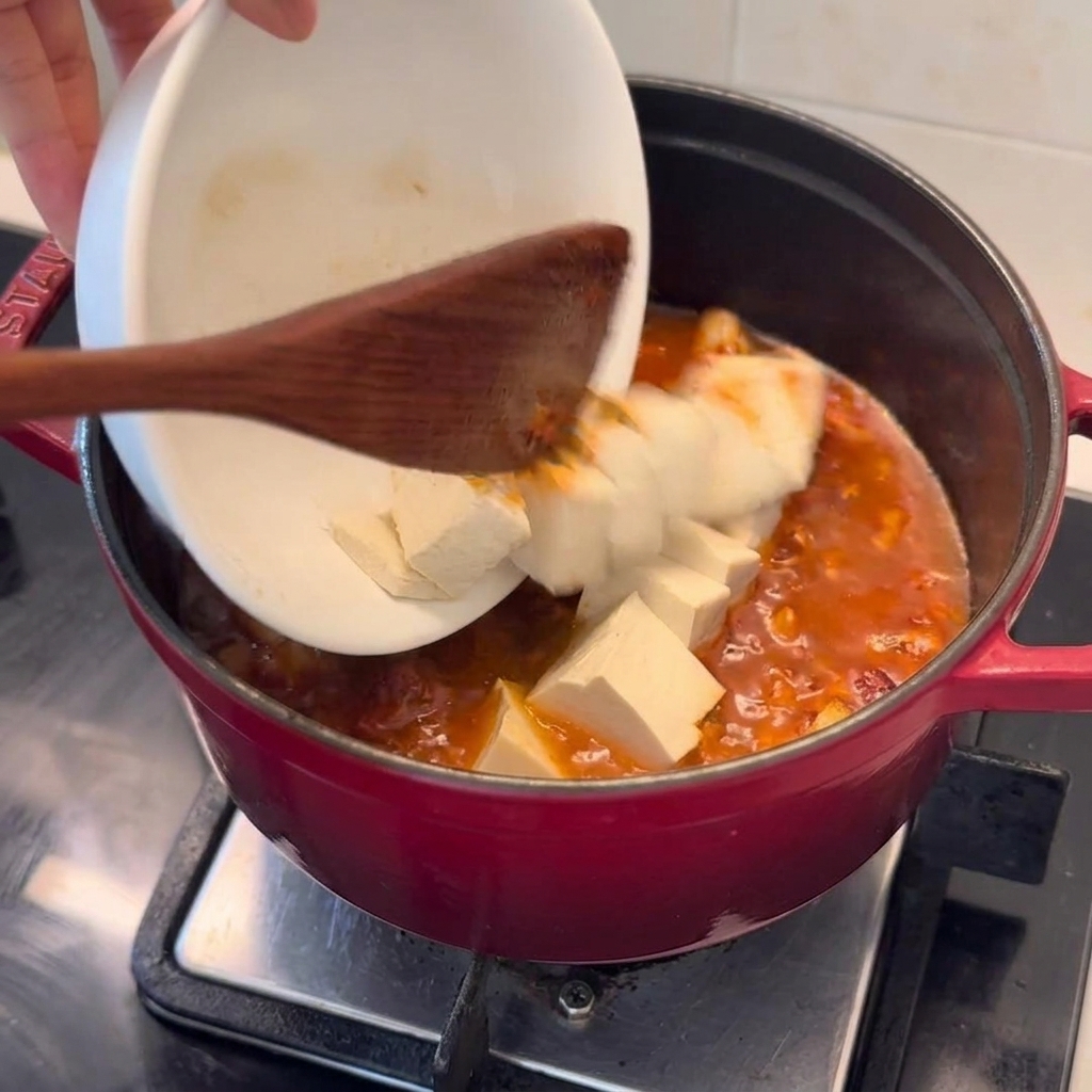 Cubos de tofu fresco vertiéndose con cuidado de un tazón blanco a una olla roja profunda con el estofado burbujeante.