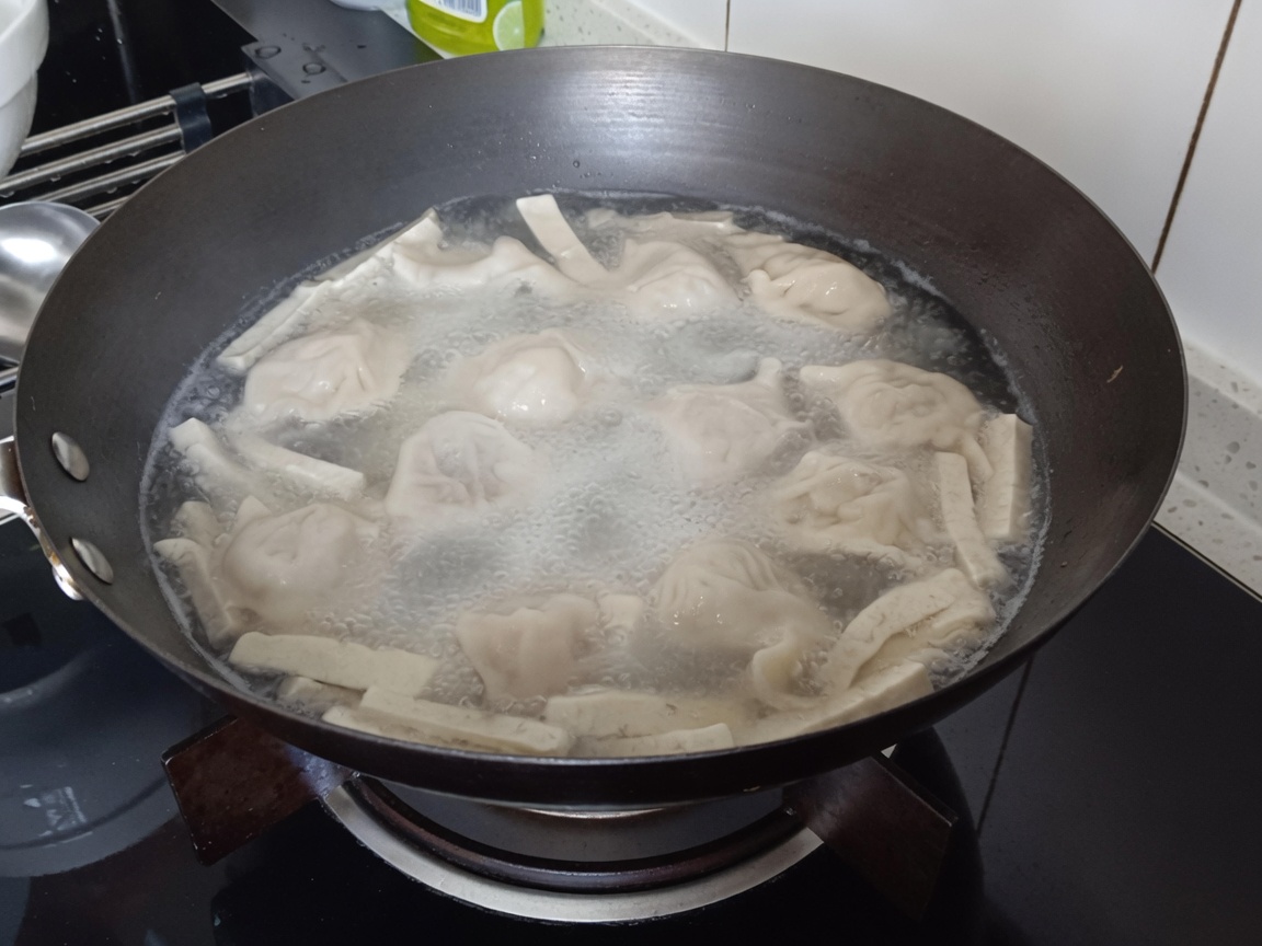 Primer plano de fideos vermicelli siendo sumergidos en la olla hirviendo con dumplings y tofu.