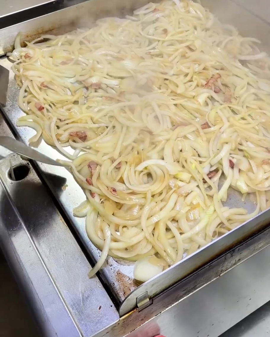 Una espátula removiendo una gran montaña de cebollas blancas ablandándose mezcladas con bacon en una plancha metálica.