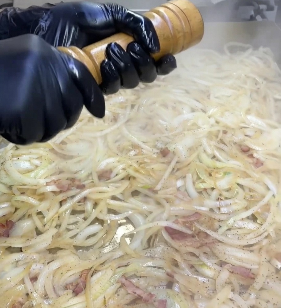 Manos con guantes negros girando un molinillo de madera sobre una plancha con cebollas y bacon.