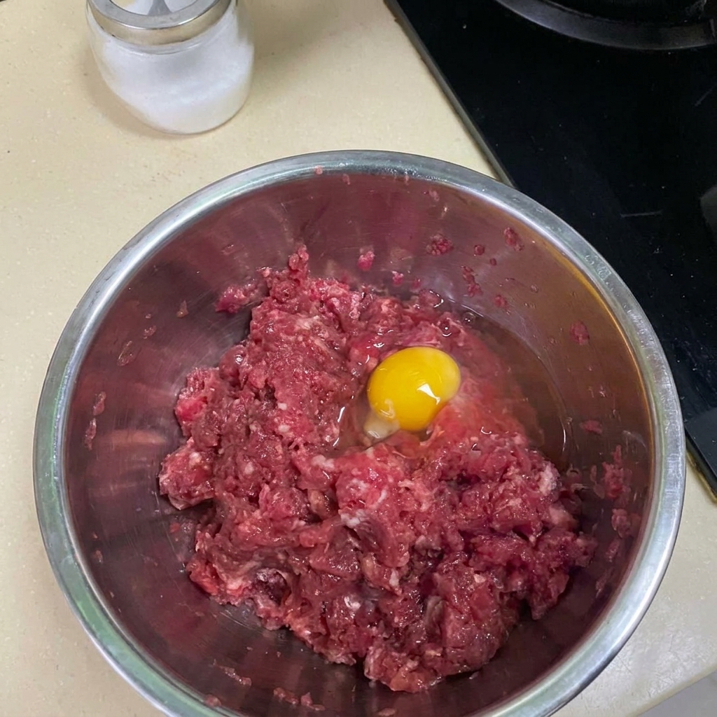 Carne picada siendo mezclada con especias y un huevo en un bol metálico.