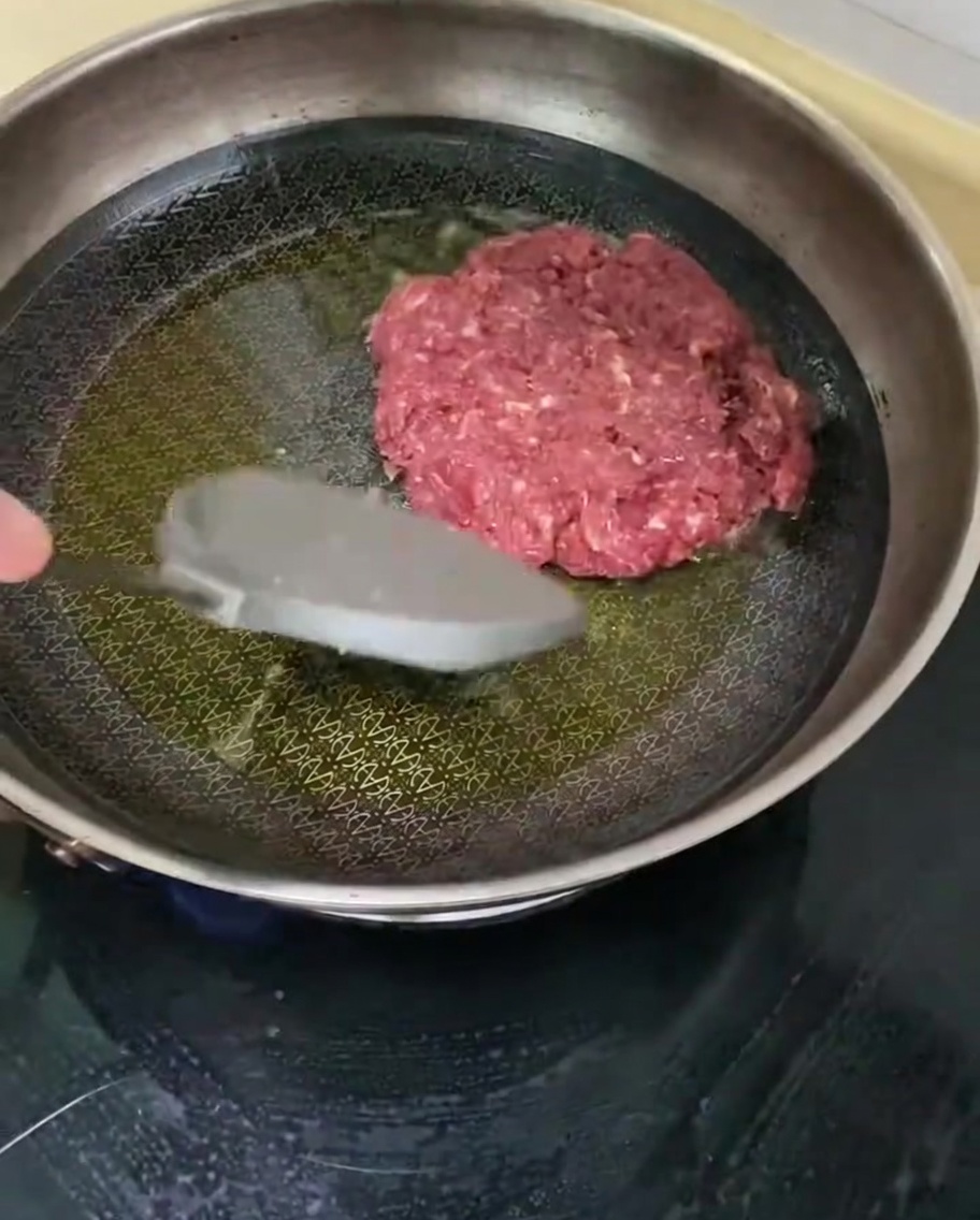 Una hamburguesa sazonada friéndose en una sartén caliente con aceite.