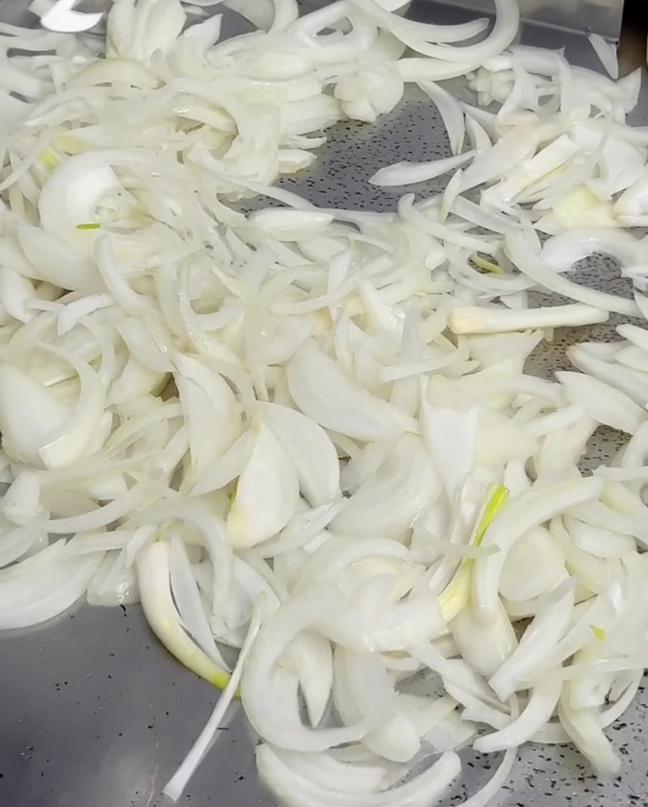 Mantequilla derretida vertiéndose desde una cacerola pequeña sobre un montón de cebollas en una plancha caliente.