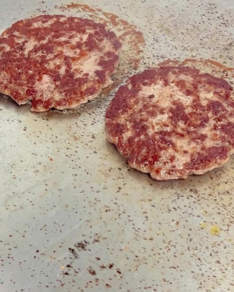 Lonchas de queso amarillo derritiéndose sobre hamburguesas de ternera calientes y doradas en una plancha.