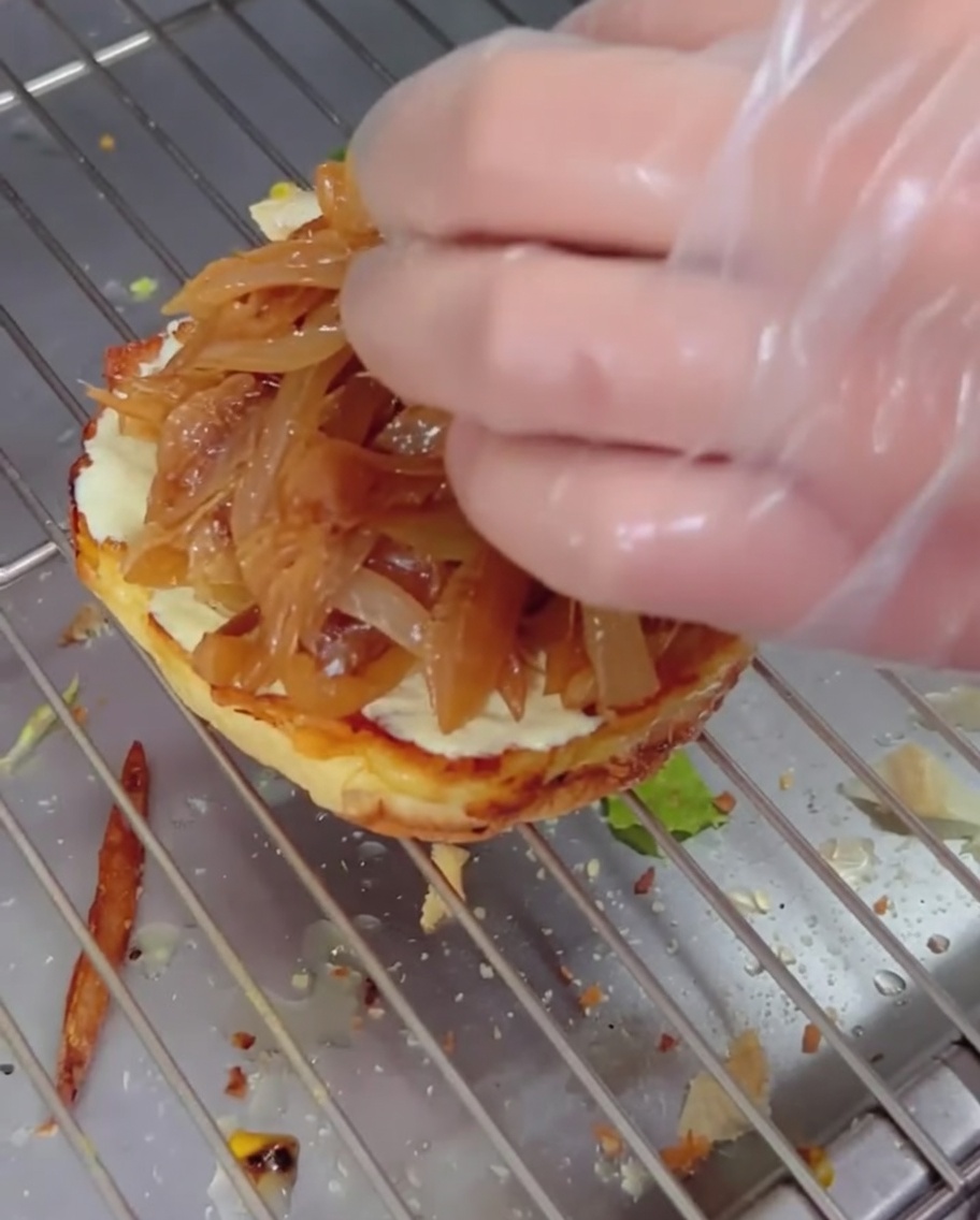 Cebollas caramelizadas siendo colocadas sobre un pan de hamburguesa con una hamburguesa con queso apilada encima.