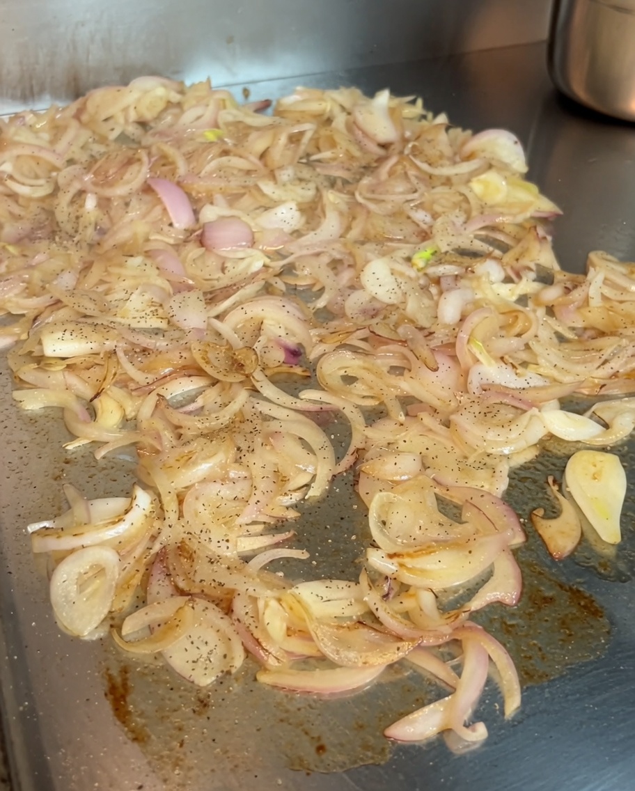 Cebollas caramelizándose en la plancha siendo sazonadas con un poco de pimienta negra y vibrante pimentón dulce húngaro.