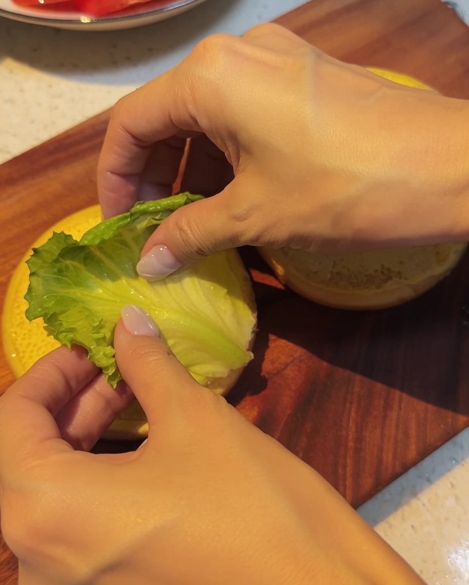Hojas de lechuga fresca siendo colocadas cuidadosamente sobre el pan inferior.