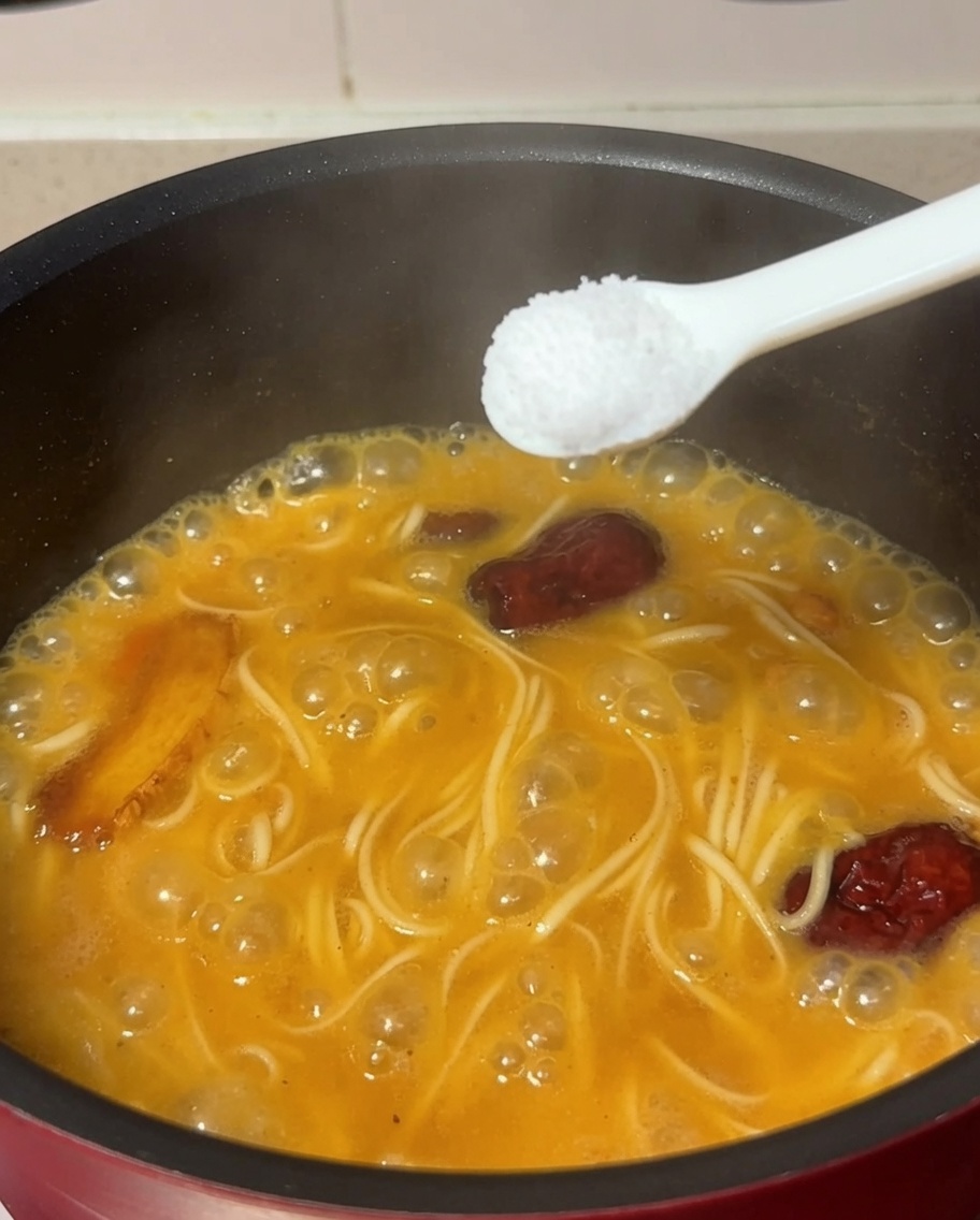 Una cucharada de sal añadiéndose a la olla que contiene los fideos y la sopa de pollo hirviendo.