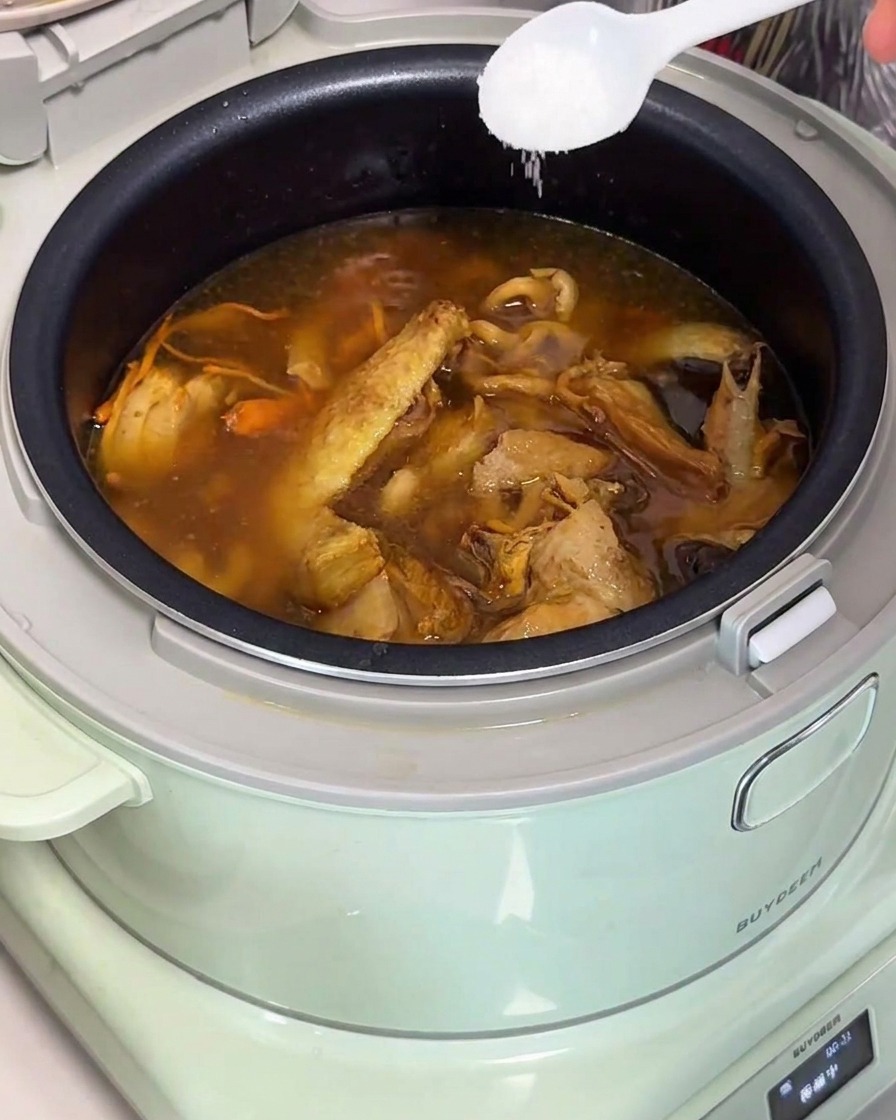 Añadiendo sal a la sopa de pollo en la olla arrocera con una cuchara.