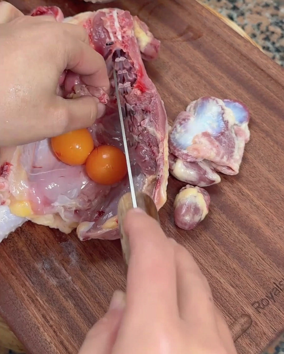 Manos usando un cuchillo para marcar las menudencias de pollo crudas junto a huevos de gallina en una tabla de madera.