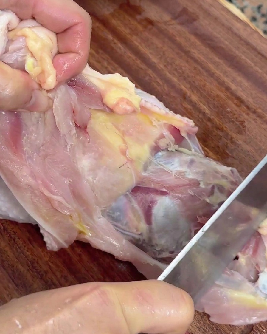 Manos sosteniendo un pollo crudo usando un cuchillo para separar la carne de la carcasa.