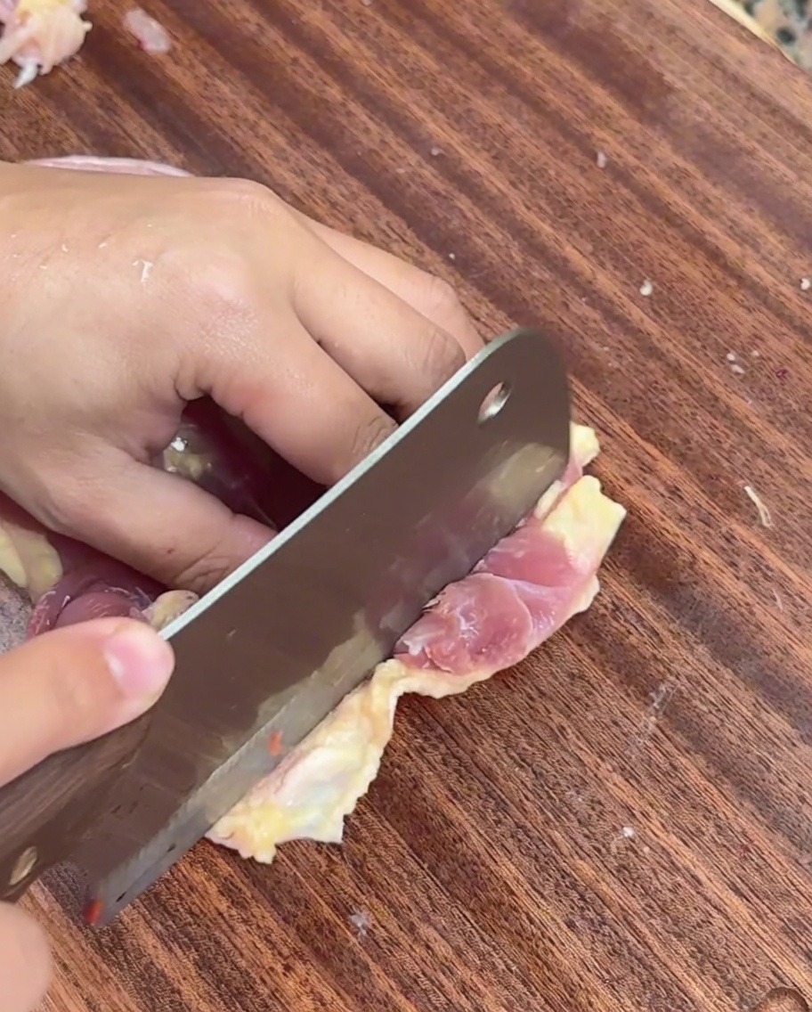 Una mano usando un cuchillo para rebanar carne de pollo deshuesada en tiras finas sobre una tabla de madera.