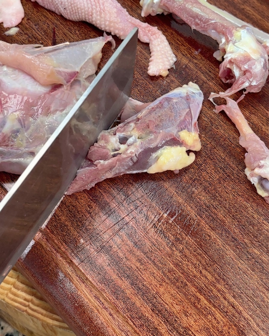 Un cuchillo troceando un esqueleto de pollo crudo en piezas grandes sobre una tabla de madera.