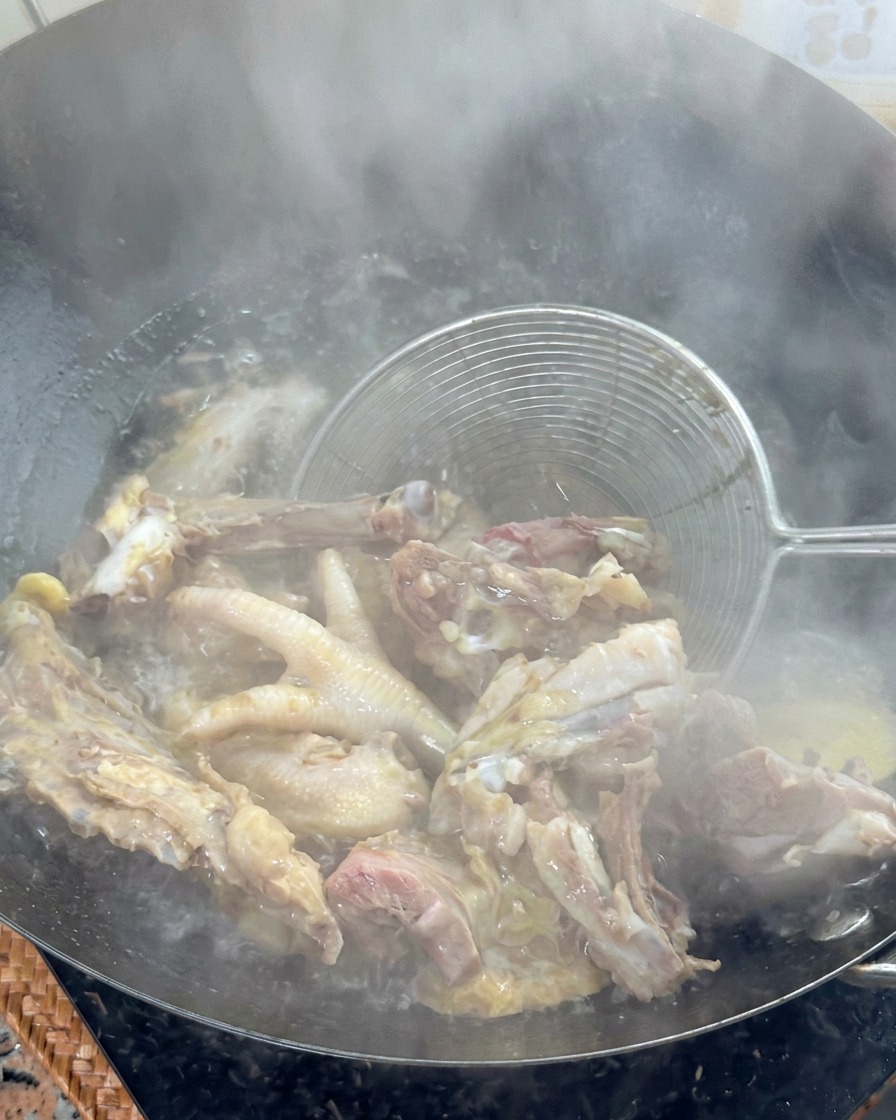 Un colador de metal levantando huesos de pollo blanqueados de un wok con agua hirviendo.